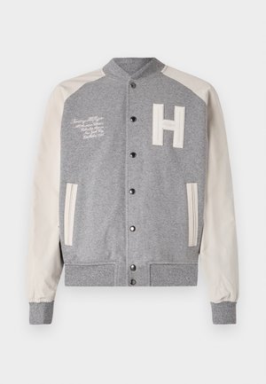 Jachetă de tip varsity gri cu mâneci din piele ecologică bej. Are un patch cu litera "H", text brodat, nasturi tip snap și manșete și tiv cu nervuri.