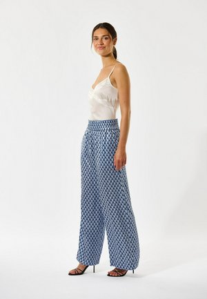 Ženska, oblečena v belo satenasto camisolo, modre široke hlače z vzorcem in črne sandale z remenčki, stoji bočno in se smehlja na enobarvnem ozadju.