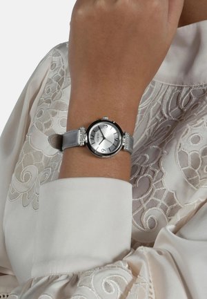 Srebrna ura s okroglo črno obrobo, teksturirano srebrno številčnico in satensko sivo zapestnico z okrasnimi kristali. V ozadju podrobna vezenina.