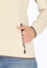 Beige fleecejas met een ritszak opzij. De binnenband is grijs. Textuurstof en een relaxte pasvorm. Hand in de zak geplaatst.
