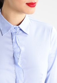 Ljusblå skjorta med knappar, rynkad placket, slät textur och spetsig krage. Framsidan visar delvis kragen och detaljerna på placket.