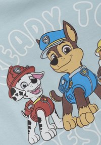 Grafika przedstawia dwa rysunkowe psy na jasnoniebieskim tle. Jeden pies jest brązowy i ma niebieski mundur policjanta; drugi jest biały w czarne plamy i nosi czerwony strój strażaka.