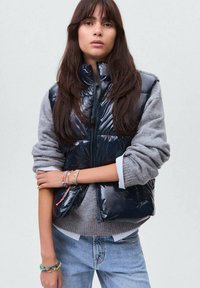 Pufferväst i glänsande svart material med hög krage, bärs över en grå tröja. Har dragkedjestängning och sidofickor. Avslappnad passform.