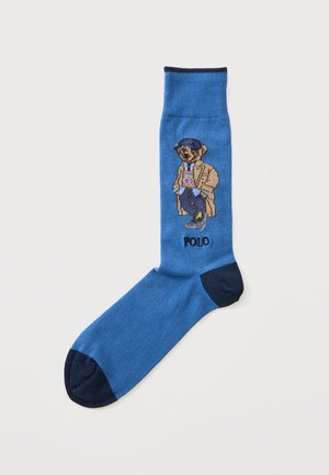 Chaussette bleue avec bout et talon foncés, représentant un ours portant un trench, une casquette et des baskets avec le mot "POLO" en dessous.