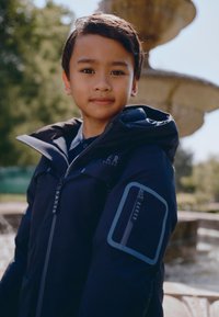 Marineblå vandtæt jakke med hætte, med kontrastfarvede lynlåse, logoaccenter og en lynlåslomme på venstre ærme.