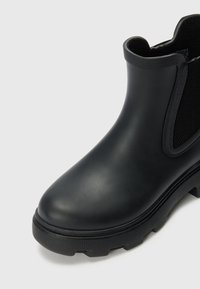 Black rubber Chelsea boot with textured side panels, rounded toe, and a chunky, patterned sole. Smooth, matte finish on the upper.