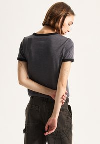 Grå kortærmet t-shirt med sort kant, der har en rund halsudskæring og en afslappet pasform, parret med sorte jeans med et edderkoppespindsmønster.