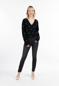 Cardigan nero con scollatura a V, decorato con motivi a cuore dorati. Indossato sopra jeans scuri e tacchi alti color crema.
