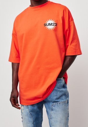 Orange cotton oversized t-shirt with short sleeves. Features a black sun graphic with "SUM23" text on the left chest.
