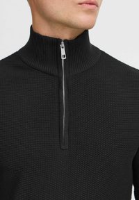 Pull-over noir à demi-zip, avec un motif en tricot texturé, col haut et détail de fermeture éclair argentée. Tissu doux et coupe ajustée.