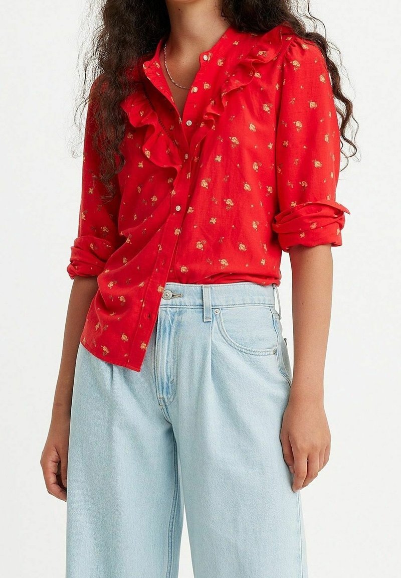 Chemise rouge à boutons avec col à volants, ornée de petites impressions florales. Associée à un jean large bleu clair.