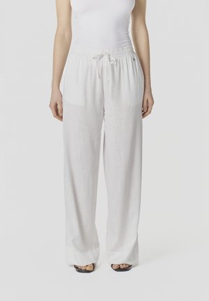 Kobieta nosząca luźne białe lniane spodnie z elastycznym pasem i sznurkiem do regulacji, połączone z czarnymi sandałami na otwartym palcu.