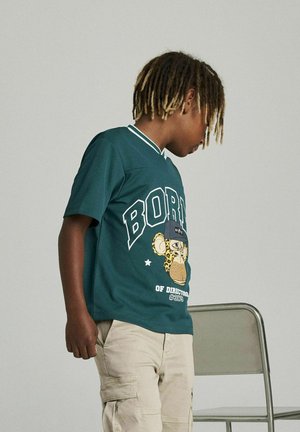 Grön kortärmad skjorta med en grafisk design av en apa, kontrasterande text och en vit krage. Beige cargoshorts och en stol synliga.