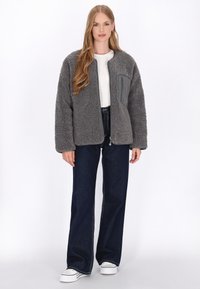 Graue Fleecejacke mit einer Reißverschlusstasche auf der Brust, kombiniert mit dunkelblauen weit geschnittenen Jeans und weißen Sneakers. Weicher Stoff und entspannter Schnitt.