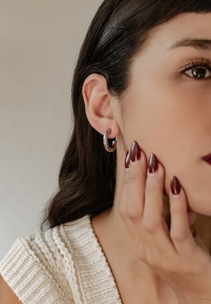 Silberne Creolen an dem Ohr eines Modells, die eine polierte Oberfläche zeigen. Das Modell hat glattes, dunkles Haar und tiefrote, manikürte Nägel.