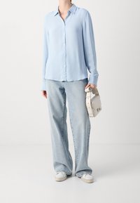 Blusa azzurro chiaro a maniche lunghe con colletto e chiusura a bottoni, abbinata a jeans a vita alta di un blu chiaro e sneakers bianche. Porta una borsa color crema.