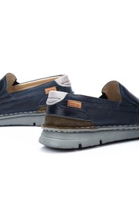 Zapatillas deslizantes de cuero azul marino con suela de goma gris, con detalles en gamuza marrón y un pequeño parche con el logo en el talón.