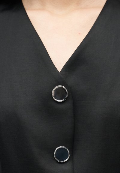 Μαύρο ρούχο με V-λαιμόκοψη και δύο μεγάλα λαμπερά μαύρα κουμπιά με ασημένιες περιφέρειες, που αφήνει να φαίνεται το δέρμα στο πάνω μέρος του στήθους πάνω από το λαιμόκοψο.