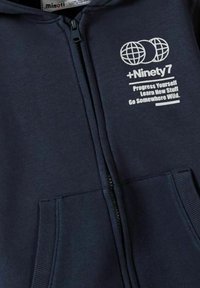Námornícka modrá mikina na zips z mäkkého materiálu, ktorá má predné vrecko a biele potlačený text a dizajn globusu na ľavej strane hrude.