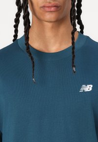 Turkio spalvos medvilninis marškinėliai su apvalia iškirpte, dekoruoti mažyte baltos spalvos "New Balance" logo kairėje krūtinės pusėje ir sklandžiu paviršiumi.