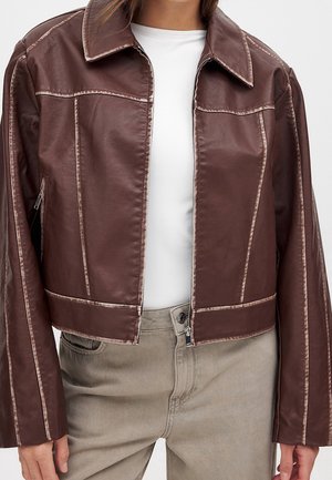 Mujer con chaqueta corta de cuero marrón con costuras visibles sobre una camisa blanca y pantalones beige.