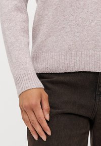 Suéter tricotado rosa claro com punhos e barra canelados, combinado com jeans castanhos escuros, destacando a textura e o contraste de cores.