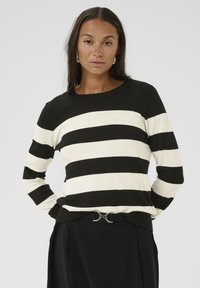 Pull rayé noir et crème avec des manches longues et un col rond. Texture de tricot fine, associé à une ceinture noire avec un accent métallique.