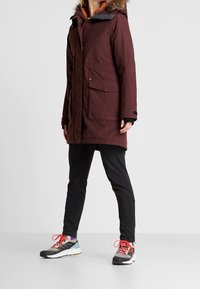 Person som bär en lång mörk vinröd vinterrock med pälsluva, svarta smala byxor och grå sneakers med klara orangea snören, som står på ett vitt golv.