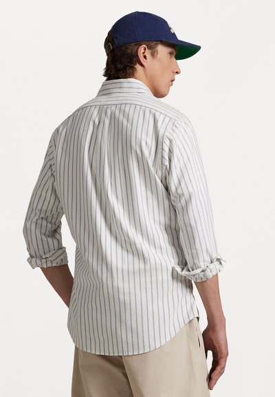 Striped shirt in white and light blue, rolled sleeves, curved hemline, soft fabric. Worn with beige pants, viewed from the back.