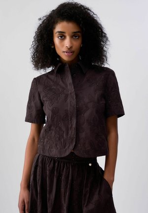 Donna con capelli ricci che indossa una camicia a maniche corte marrone scuro con bottoni e una gonna abbinata con ricami floreali, in piedi davanti a uno sfondo semplice.