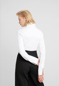 Chemise blanche à boutons avec un col structuré et des manches longues, portée avec un pantalon noir taille haute. Présente des accents contrastés aux poignets.