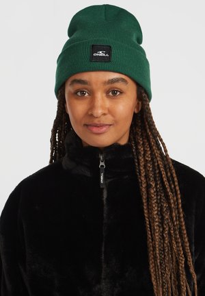 Green knitted beanie with a cuff and a black brand patch. Worn with a black fleece jacket featuring a zip front and high collar.