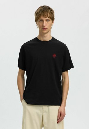 Młody mężczyzna o krótkich włosach, ubrany w czarną koszulkę z małą czerwoną grafiką na piersi i jasno beżowe spodnie, stojący prosto.