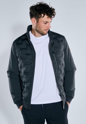 Black padded jacket with high collar, zip closure, and side pockets. Worn over a white T-shirt, featuring a quilted texture.