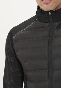 Mustavärinen toppaliivi korkealla kauluksella, jossa on kiiltävät yksityiskohdat ja tyylikäs vetoketju. Sisäkerros koostuu istuvasta, tekstuuripintaisesta mustasta pitkähihaisesta paidasta.