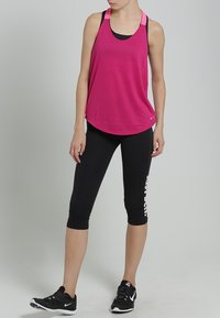 Femme debout portant un débardeur rose vif, des leggings courts noirs avec du texte blanc, et des chaussures de sport noires sur un fond gris.