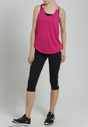 Kobieta stojąca, ubrana w jaskrawo różowy, bezrękawnik, czarne legginsy do połowy łydki z białym napisem oraz czarne sportowe buty na szarym tle.