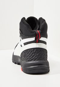 Valkoinen ja musta puolikorkea saapas, jossa on teksturoitu musta kumipohja, pehmustettu kaulus ja punainen yksityiskohta kantapään lähellä. Varustettu hengittävällä verkkomateriaalilla.