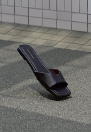 Dunkelbraune offene Sandale mit quadratischer Spitze und breitem Riemen, liegend auf grauem Asphalt neben gefliestem Bürgersteig.