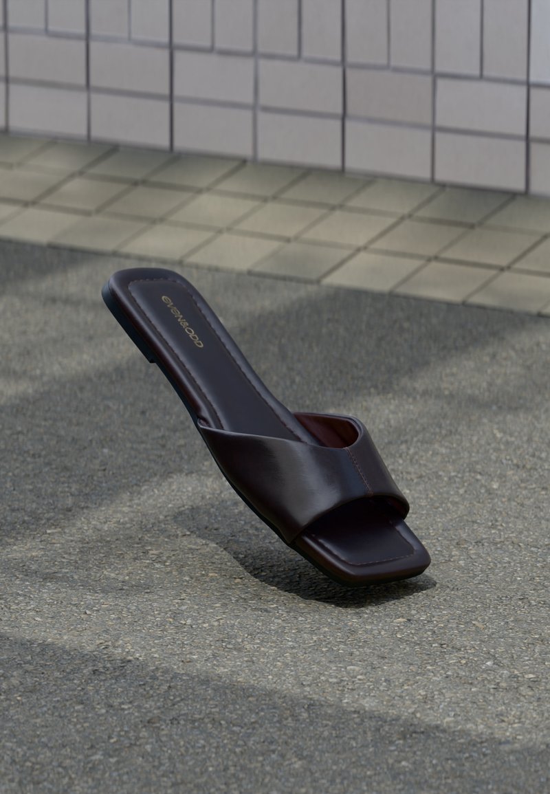 Sandale slide marron foncé à bout carré et large bride, posée sur un goudron gris près d'un trottoir carrelé.