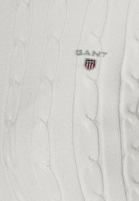 Pullover bianco a maglia grossa con un piccolo logo ricamato "GANT" e una toppa a forma di scudo rossa e blu sul petto.