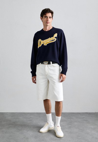 Laevastiku crewneck sviiter tumesinisest kangast kollase "Dsquared²" logo'ga, koos valgete teksashortside, valgete sokkide ja valgete tennistega, seistes hallil põrandal.