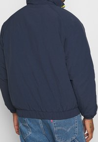 Navyblauwe donsjack met een hoge kraag, elastische manchetten en een zoom; gladde textuur met een lichte glans en opvallende stiksel details.