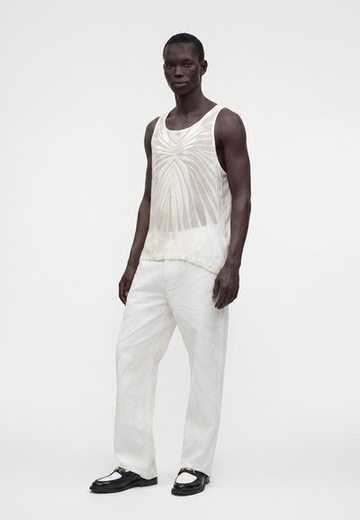 Homme à la peau foncée portant un haut sans manches blanc à motifs, un pantalon blanc ample et des mocassins noirs et blancs, debout devant un fond uni.
