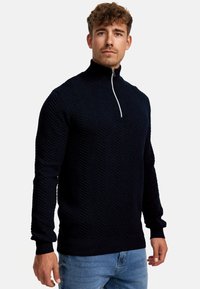 Marineblauer Zip-Neck-Pullover mit strukturiertem Fischgrätmuster. Ribbed Bündchen und Saum. Das Modell trägt helle Jeans und steht vor einem schlichten Hintergrund.