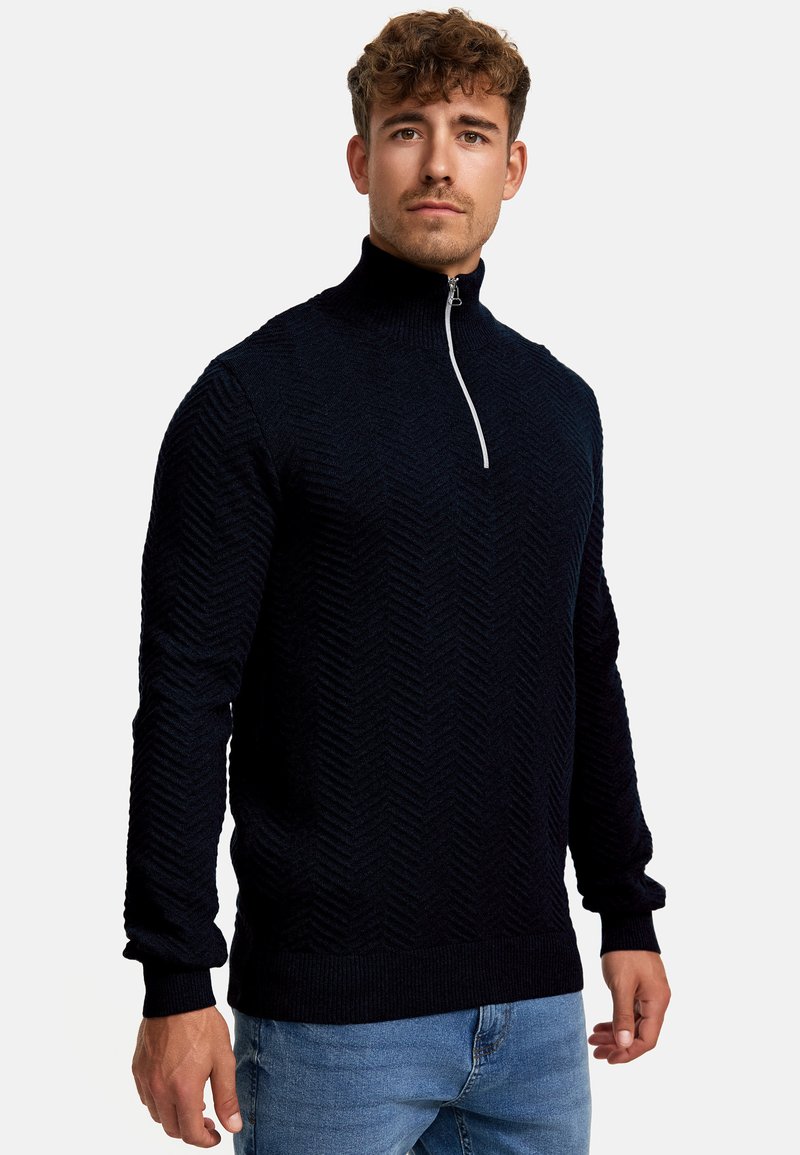 Marineblauer Zip-Neck-Pullover mit strukturiertem Fischgrätmuster. Ribbed Bündchen und Saum. Das Modell trägt helle Jeans und steht vor einem schlichten Hintergrund.