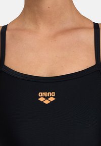 Maillot de bain noir au tissu lisse, avec de fines bretelles et un logo "arena" orange au-dessus d'un petit design en triangle.