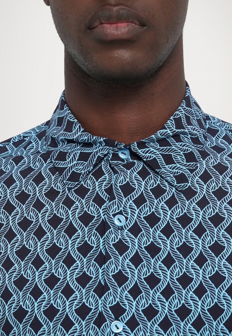 Acercamiento de una persona con una camisa azul marino con un patrón de cuerdas azul claro y botones azules a juego, mostrando el cuello y la parte superior del pecho.