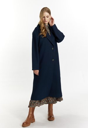 Cappotto di lana blu navy con ampio colletto, bottoni frontali e maniche lunghe, indossato sopra un vestito floreale con volant e stivaletti marroni alla caviglia.