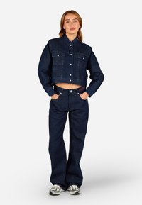 Σκούρο μπλε τζιν μπουφάν με κοντό σχέδιο, μπροστινές τσέπες και κλείσιμο με κουμπιά; συνδυασμένο με ταιριαστά τζιν παντελόνια με ψηλή μέση.